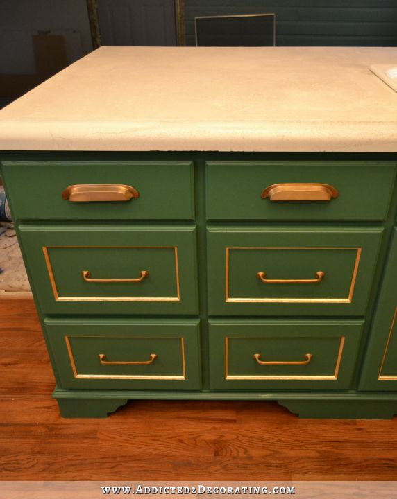 kitchen peninsula cabinets painted green with gold leaf accents and antique brass hardware, with white concrete countertop