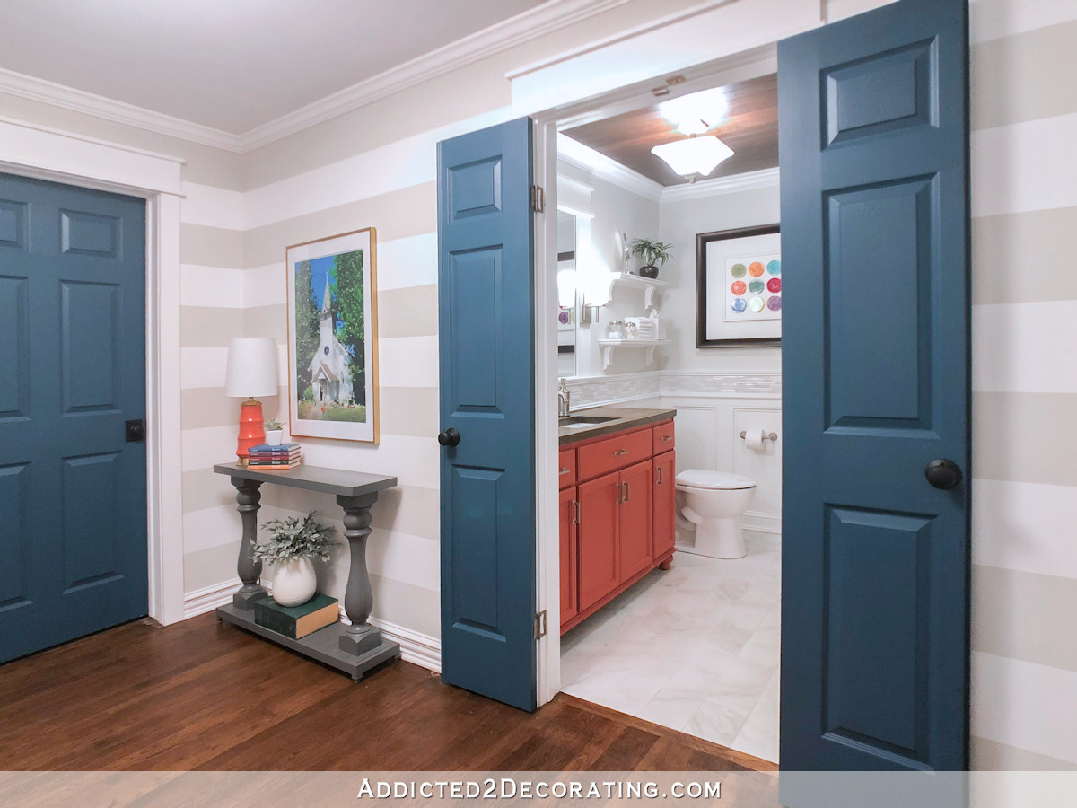 Hallway with horizontal striped walls in gray and white, with dark teal interior doors. Bathroom with coral vanity and colorful artwork.