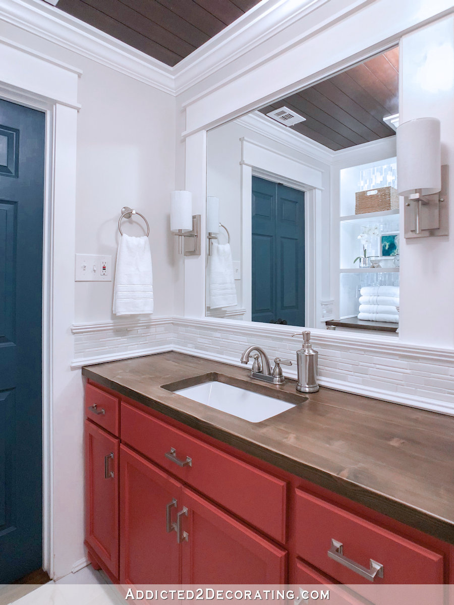 Coral bathroom vanity with stained wood countertop, dark teal bathroom doors, white mosaic tile backsplash.