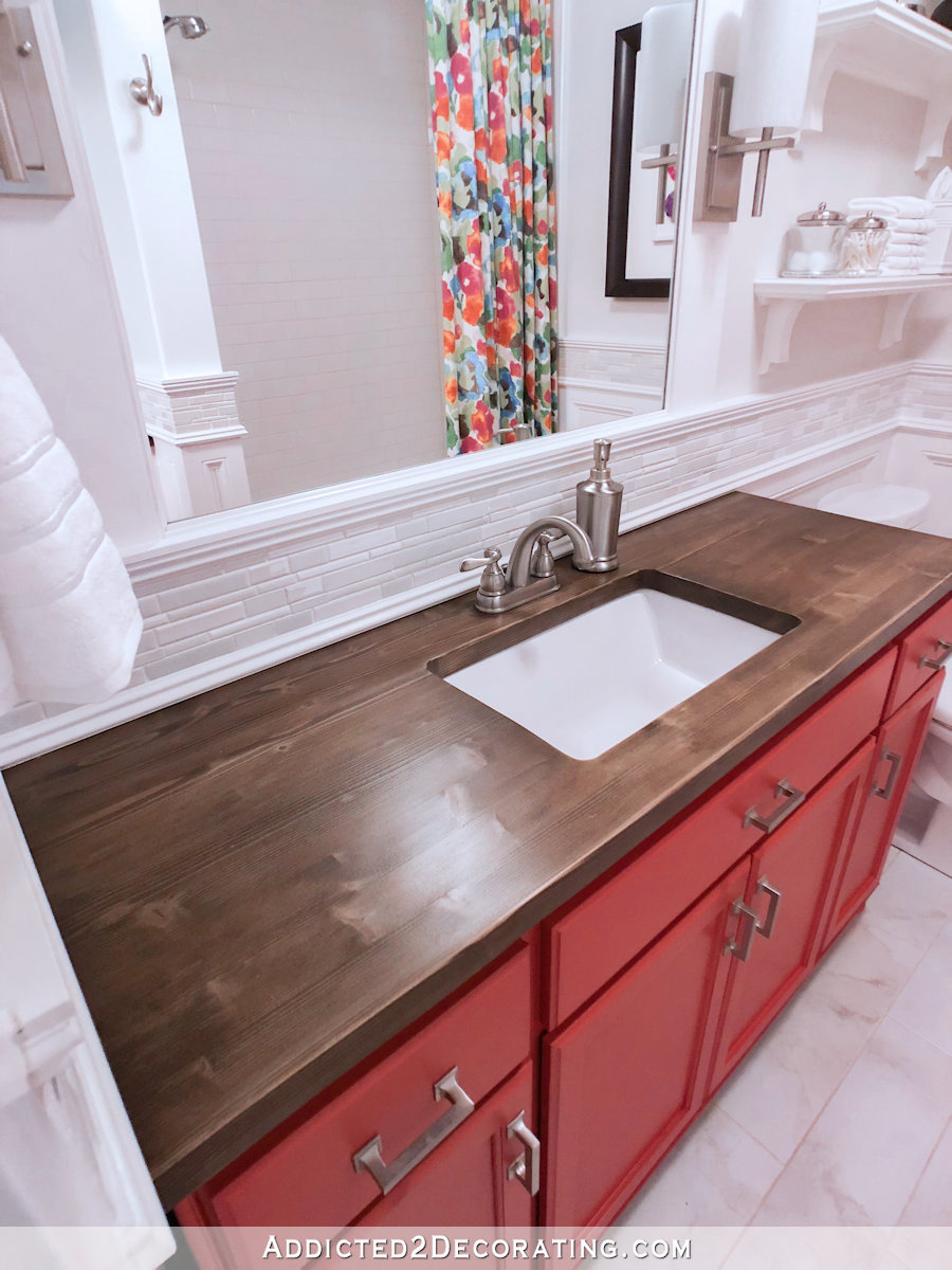 Coral vanity in bathroom with wood countertop and undermount sink, white mosaic tile backsplash, bright floral shower curtain.