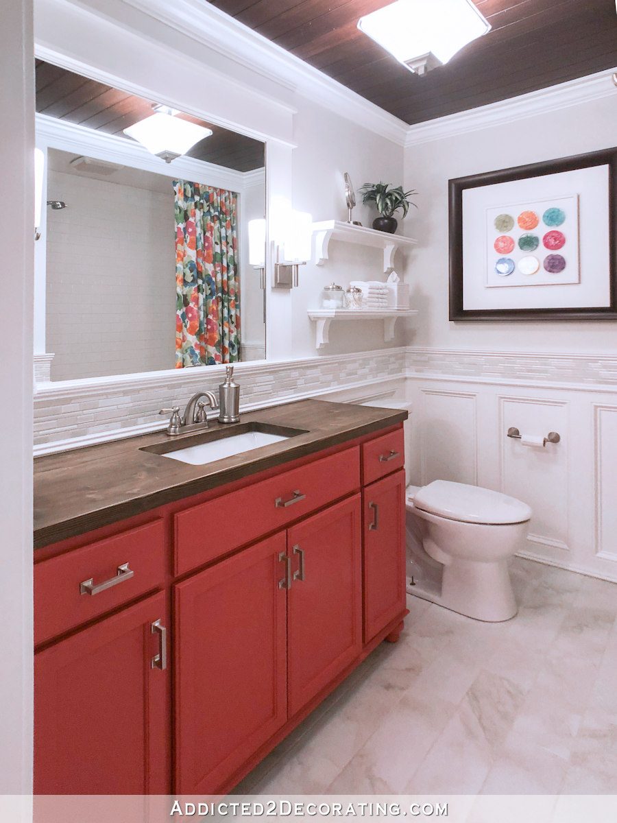 Colorful small bathroom makeover with coral vanity, wood countertop, white mosaic tile accents, and floral shower curtain.