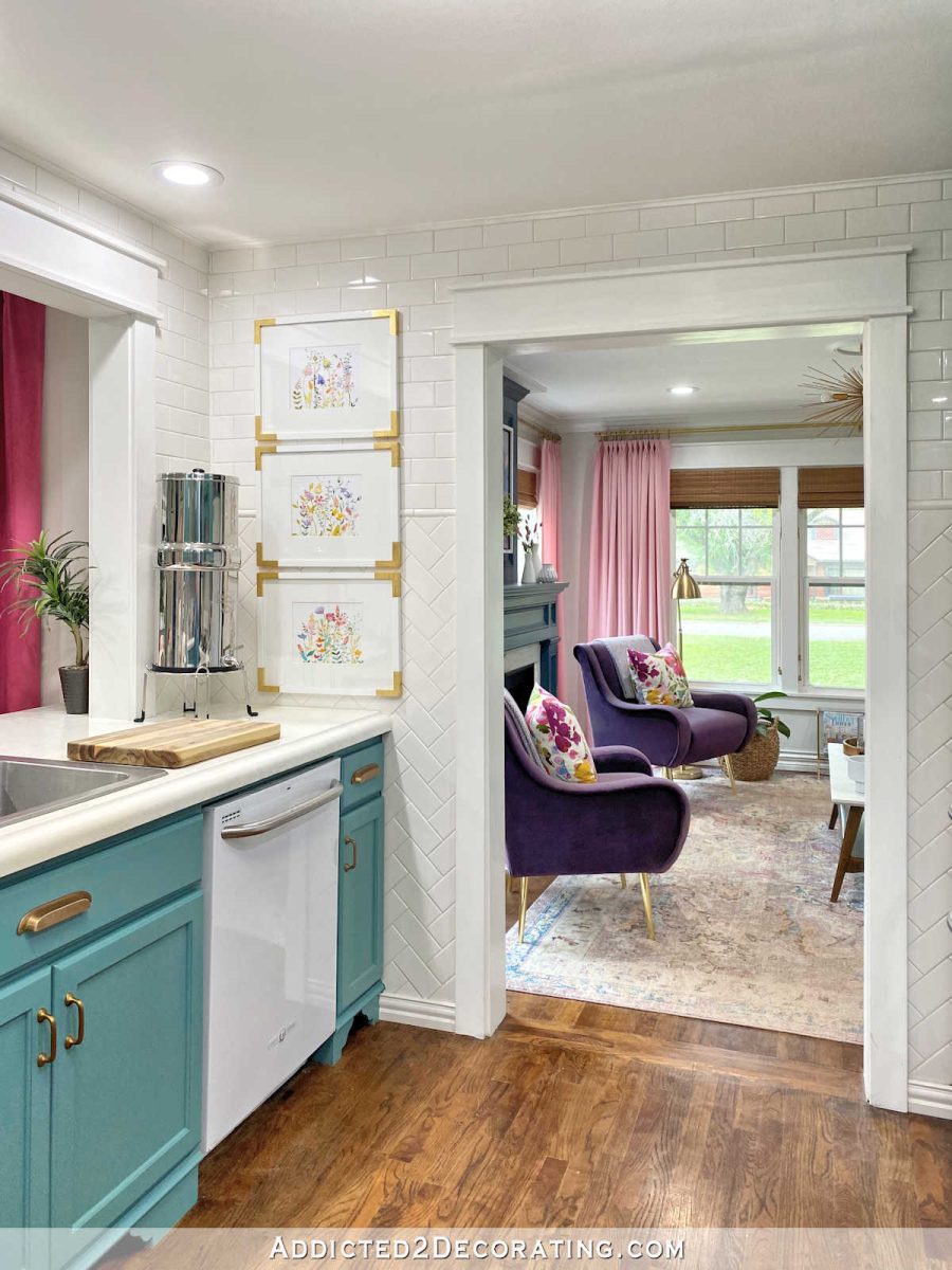 kitchen with teal cabinets and white subway tile walls, cased opening into living room with pink curtains, purple chairs, teal fireplace