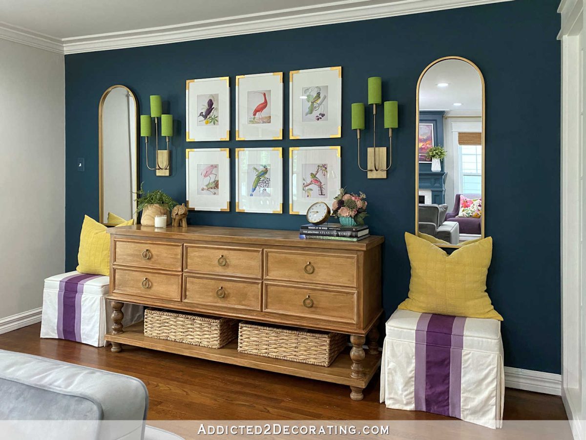 Entryway with dark teal wall, gold framed arch mirrors, grid gallery wall of bird illustrations, wall sconces, DIY credenza, and DIY upholstered ottomans