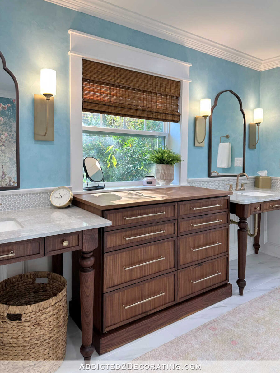 walnut storage cabinet under window flanked by two table-style vanities in a master bathroom