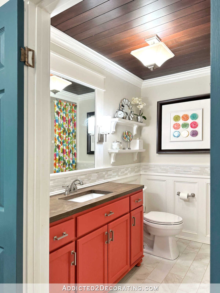 Colorful guest bathroom with white wainscots, light gray walls, floral shower curtain, wooden countertops and coral orange vanity.