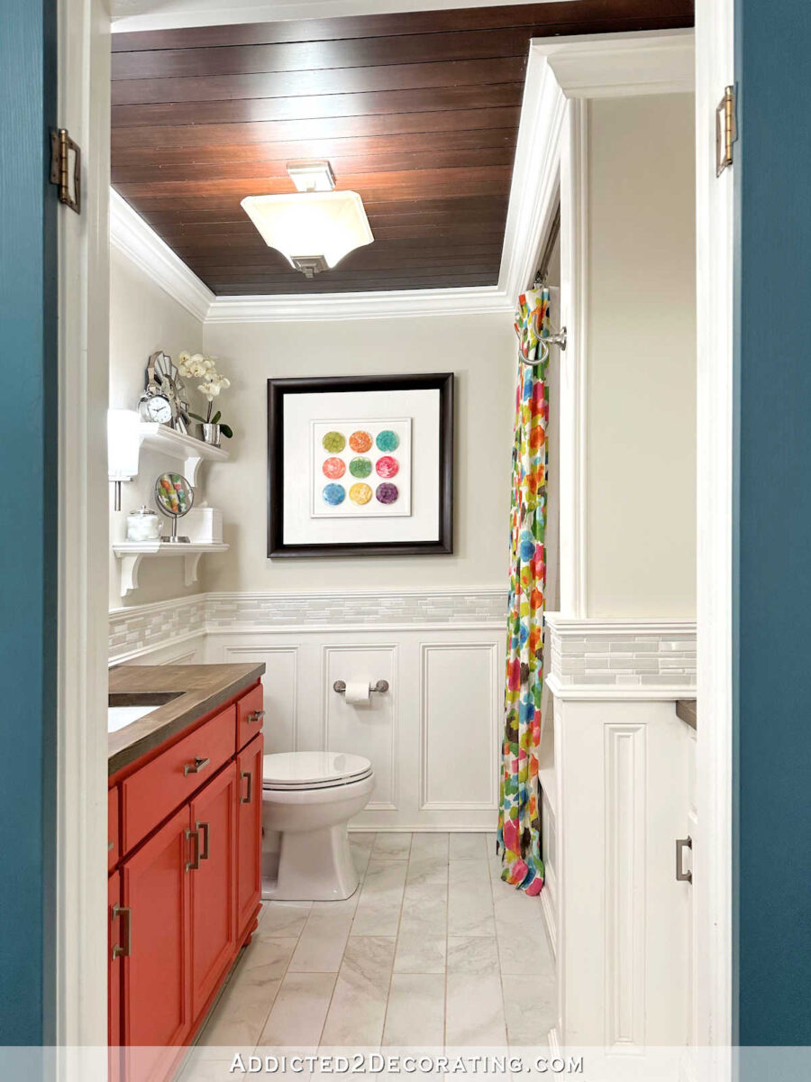 Colorful guest bathroom with stained wood ceiling, white trim and wainscoting judge's paneling, tile accent, colorful shower curtain, wood countertop, and coral vanity