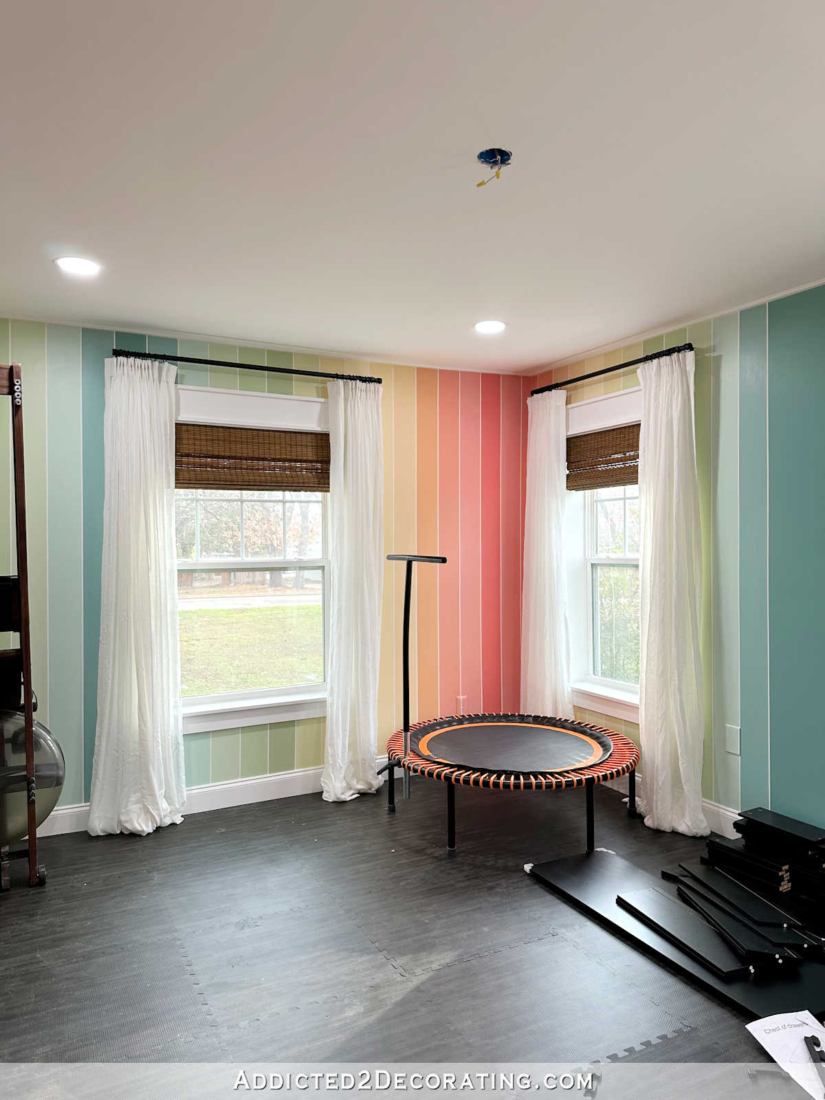 Home gym with colorful striped walls, white sheer curtains, black foam floor, woven wood shades, Bellicon rebounder