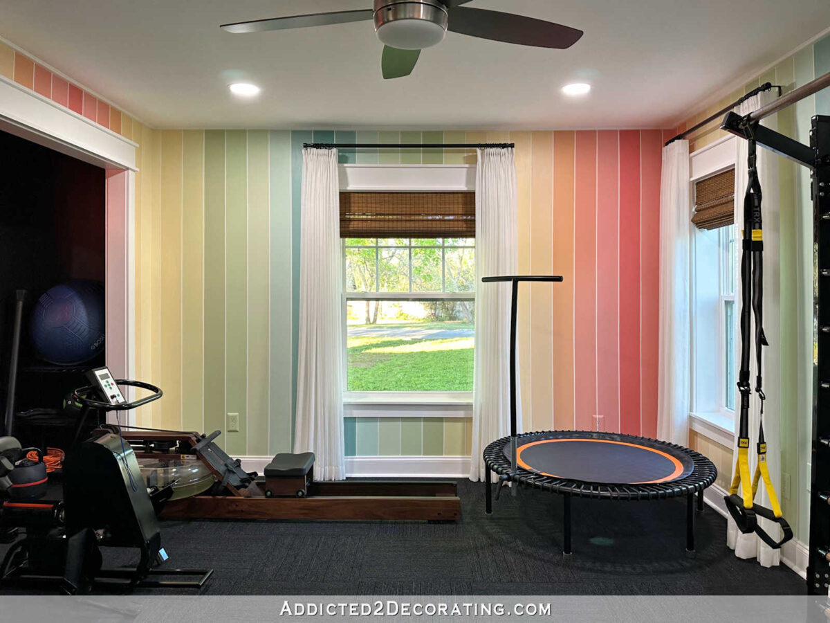 finished home gym with colorful striped walls, white sheer curtains, black carpet tiles; Bellicon rebounder, TRX suspension trainer, WaterRower rowing machine, Theracycle exercise bike