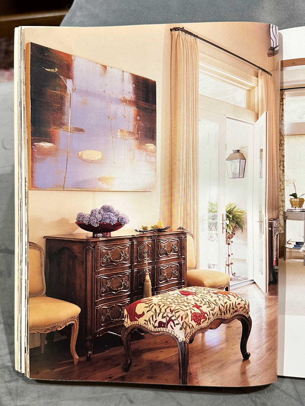 Living room with dark wood console, large painted artwork, upholstered ottoman, yellow upholstered chairs, draperies on French doors. 